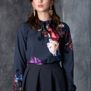 May include: A dark blue blouse with a floral print and a high neck. The blouse has a tie at the neck and long sleeves. The model is wearing a black skirt.