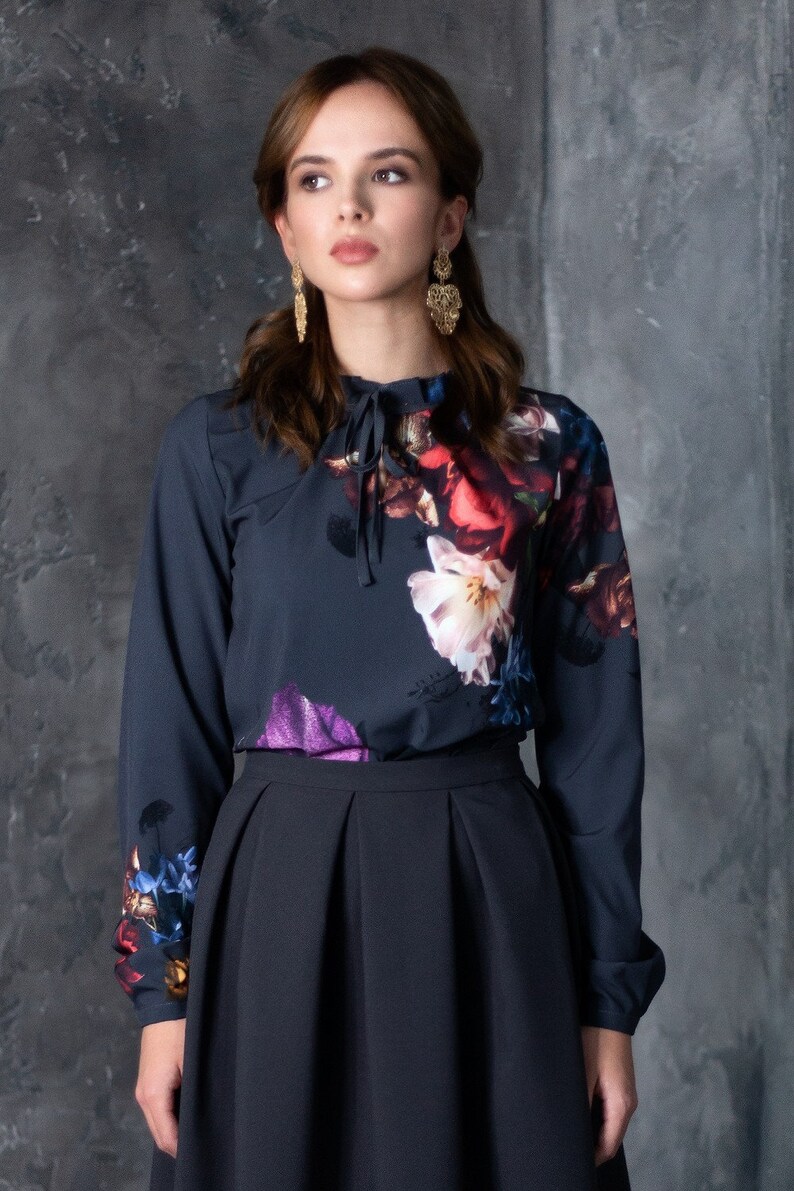 May include: A dark blue blouse with a floral print and a high neck. The blouse has a tie at the neck and long sleeves. The model is wearing a black skirt.