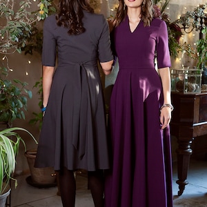 May include Two women wearing dresses. The woman on the left is wearing a charcoal gray dress with a tie at the waist. The woman on the right is wearing a deep purple dress with a v-neck.