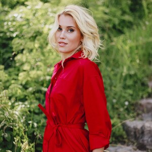 May include: A woman wearing a bright red shirt dress with a tie belt. The dress has long sleeves and a button-down front. She is wearing a white watch with a gold bezel. The background is blurred green foliage.