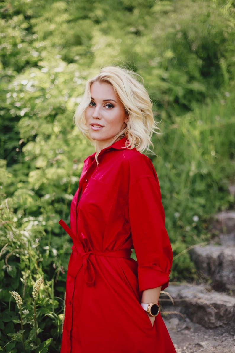 May include: A woman wearing a bright red shirt dress with a tie belt. The dress has long sleeves and a button-down front. She is wearing a white watch with a gold bezel. The background is blurred green foliage.