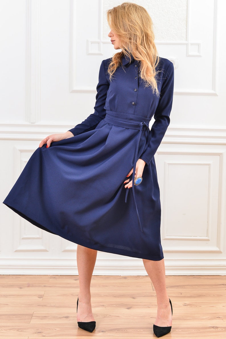 Pu&ograve; includere: Un abito midi blu navy con colletto, maniche lunghe e cintura annodata in vita.