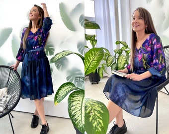 May include: A woman wearing a dark blue floral print dress with a ruffled hem. The dress has a V-neckline and 3/4 sleeves. The woman is standing in front of a white wall with green plants.