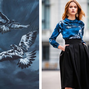 May include A blue silk blouse with a print of two black and white birds flying against a blue sky. The blouse has a white collar and long sleeves. The model is wearing a black skirt with a high waist and pleats.