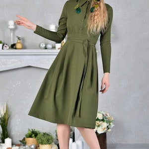 Pu&ograve; includere: Un abito lungo a maniche lunghe, di colore verde oliva, con bottoni sul davanti e una cintura a nodo. L'abito ha una gonna svasata e viene indossato con tacchi gialli.