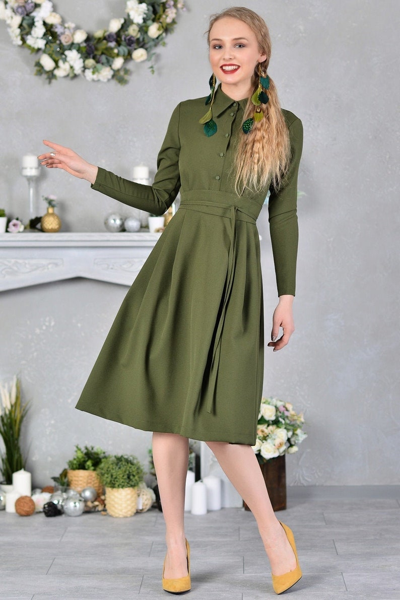 Pu&ograve; includere: Un abito lungo a maniche lunghe, di colore verde oliva, con bottoni sul davanti e una cintura a nodo. L'abito ha una gonna svasata e viene indossato con tacchi gialli.