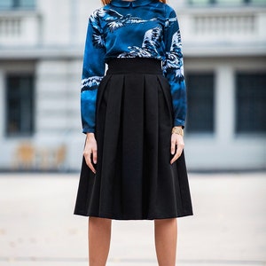 May include A woman wearing a blue and white patterned blouse with a white collar and a black midi skirt with pleats. She is wearing white heels.