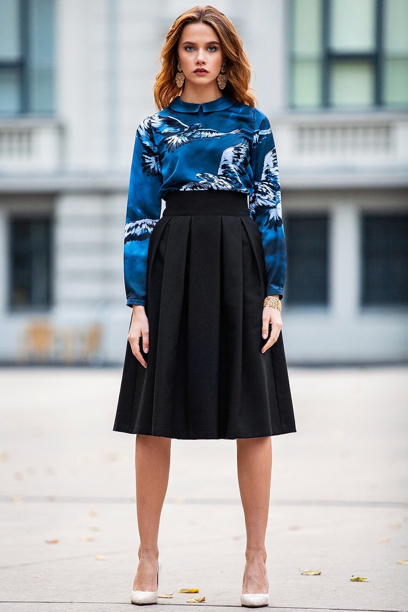 May include A woman wearing a blue and white patterned blouse with a white collar and a black midi skirt with pleats. She is wearing white heels.