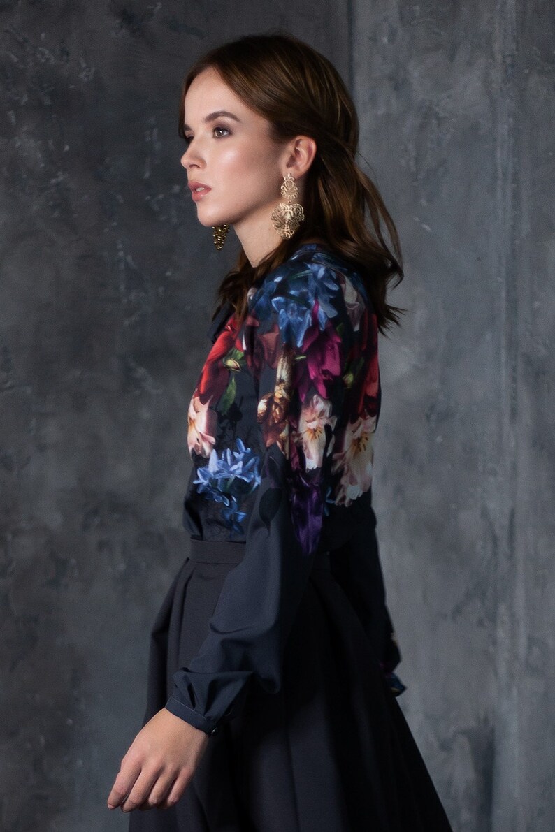 May include: A woman wearing a black long-sleeved blouse with a floral print and a black skirt. She is wearing gold earrings.