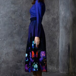 K&ouml;nnte beinhalten: Eine Frau tr&auml;gt ein blaues, lang&auml;rmeliges Kleid mit einem schwarzen G&uuml;rtel und einem Rock mit Blumenmuster. Der Rock hat einen Farbverlauf von Blau nach Lila und zeigt verschiedene Blumen in Rot-, Orange-, Blau- und Lilat&ouml;nen.