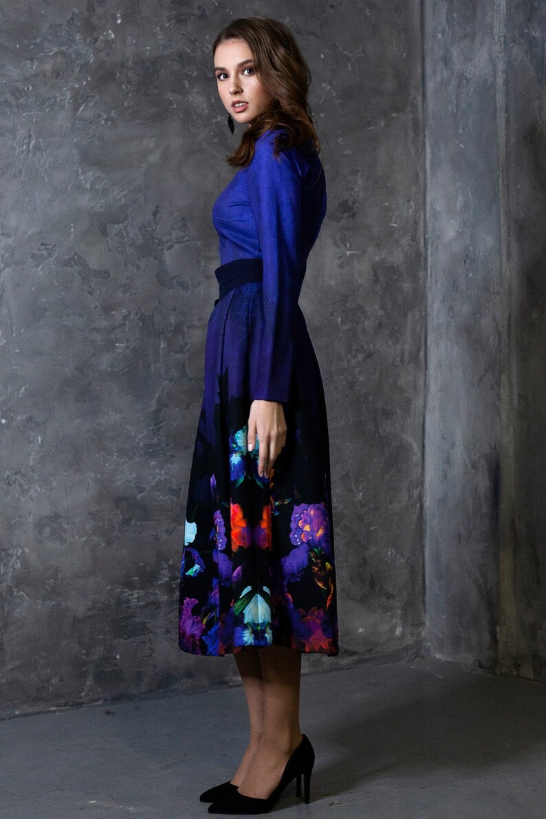 K&ouml;nnte beinhalten: Eine Frau tr&auml;gt ein blaues, lang&auml;rmeliges Kleid mit einem schwarzen G&uuml;rtel und einem Rock mit Blumenmuster. Der Rock hat einen Farbverlauf von Blau nach Lila und zeigt verschiedene Blumen in Rot-, Orange-, Blau- und Lilat&ouml;nen.