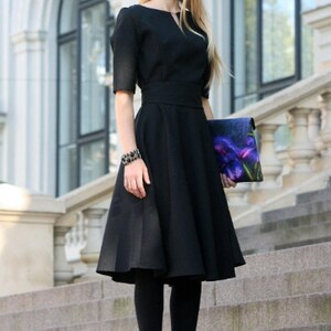 May include: A woman wearing a black dress with a fitted bodice and a full skirt. She is holding a floral clutch and wearing black tights and heels.