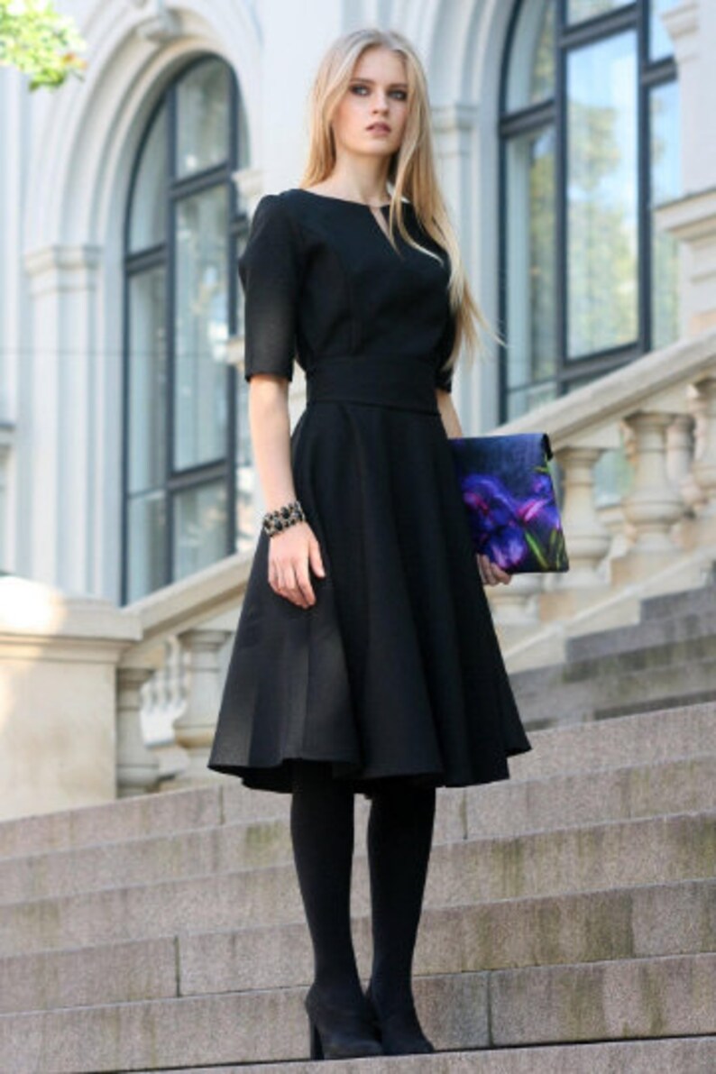 May include: A woman wearing a black dress with a fitted bodice and a full skirt. She is holding a floral clutch and wearing black tights and heels.