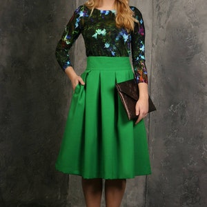 Pu&ograve; includere: Una donna indossa una gonna verde a pieghe, una camicia a maniche lunghe con stampa floreale e stivaletti marroni. Tiene una pochette marrone.