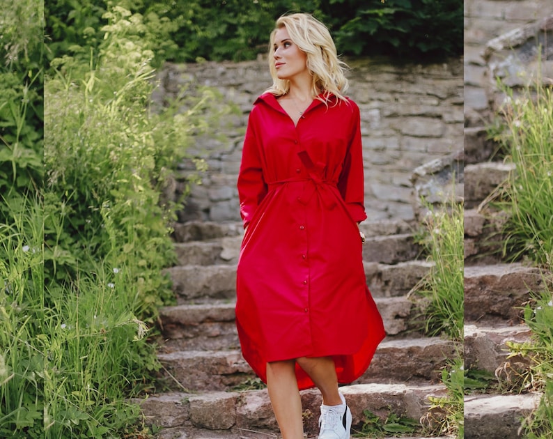 May include: A vibrant red shirt dress with long sleeves and a button-down front. The dress features a matching belt at the waist and a flowing, asymmetrical hemline. The person is wearing white sneakers. The background includes stone steps and greenery.