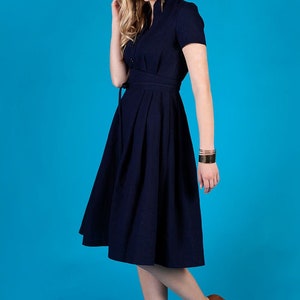 May include A navy blue midi dress with a button-up front, short sleeves, and a tie belt. The dress has a pleated skirt and is worn by a person with brown hair and brown shoes.