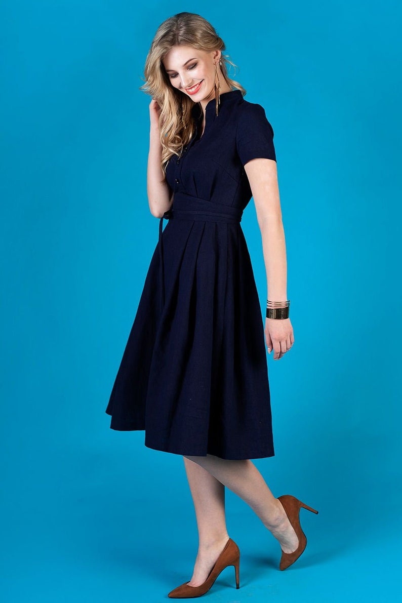 May include A navy blue midi dress with a button-up front, short sleeves, and a tie belt. The dress has a pleated skirt and is worn by a person with brown hair and brown shoes.