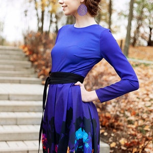 K&ouml;nnte beinhalten: Eine Frau tr&auml;gt ein lang&auml;rmeliges, violettes Kleid mit einem schwarzen G&uuml;rtel und einem Blumendruck. Das Kleid hat ein tailliertes Oberteil und einen flie&szlig;enden Rock. Sie steht auf einer Steintreppe mit Herbstlaub im Hintergrund.
