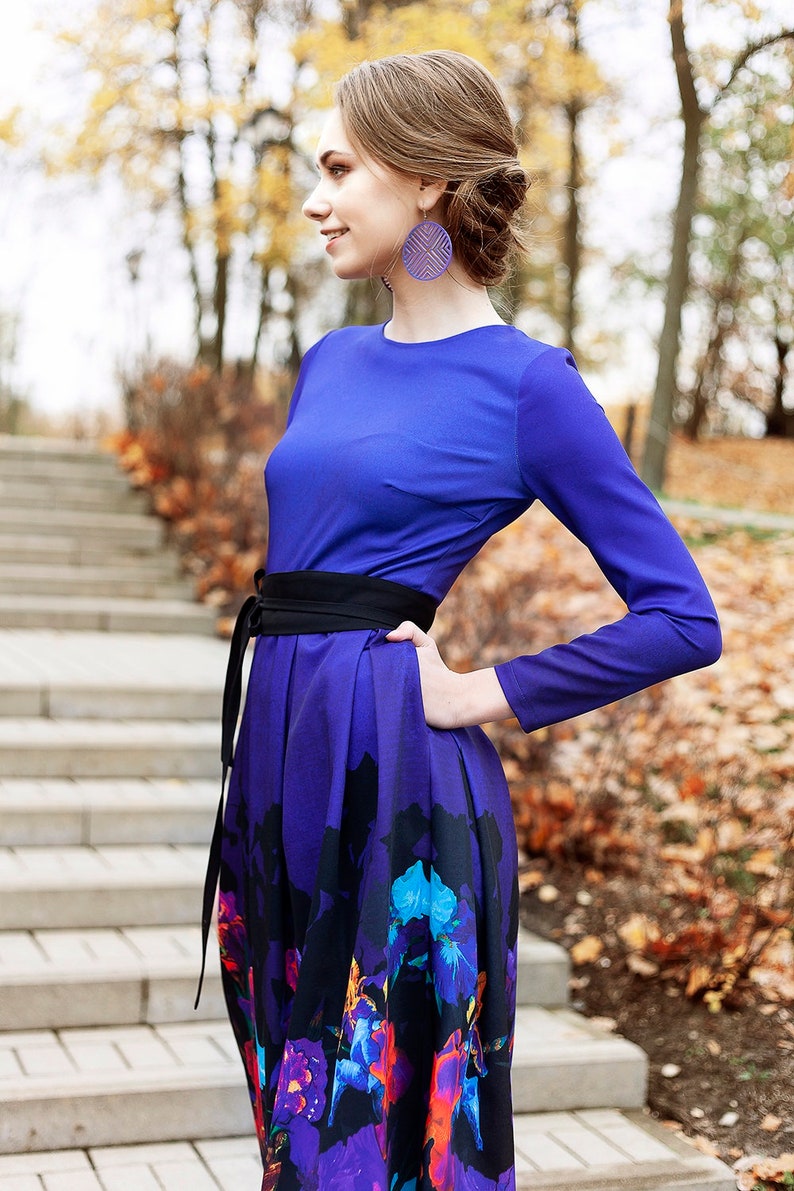 K&ouml;nnte beinhalten: Eine Frau tr&auml;gt ein lang&auml;rmeliges, violettes Kleid mit einem schwarzen G&uuml;rtel und einem Blumendruck. Das Kleid hat ein tailliertes Oberteil und einen flie&szlig;enden Rock. Sie steht auf einer Steintreppe mit Herbstlaub im Hintergrund.