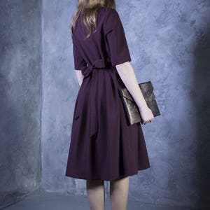 May include: A woman wearing a burgundy dress with a bow at the waist and a gold clutch.