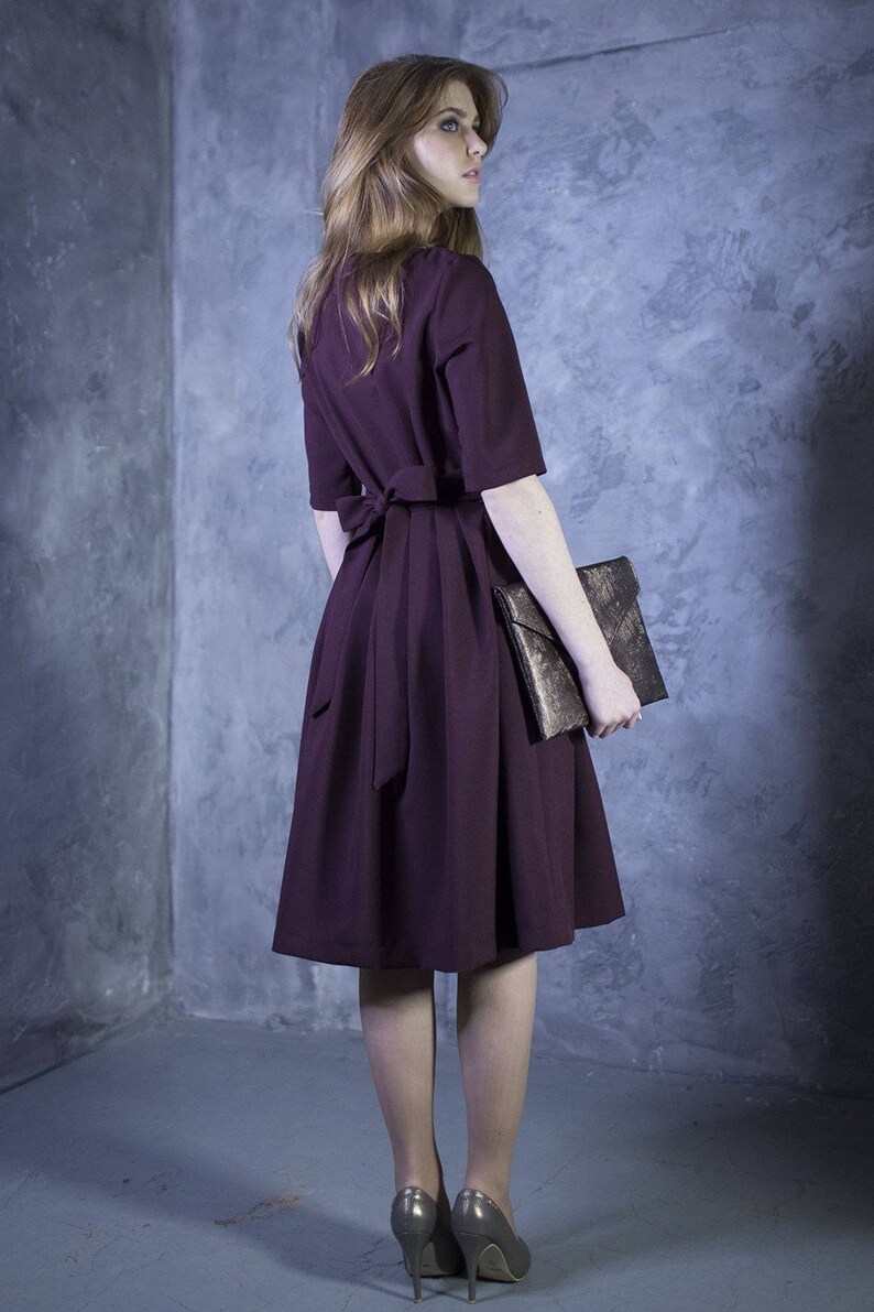 May include: A woman wearing a burgundy dress with a bow at the waist and a gold clutch.
