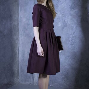 May include: A woman wearing a maroon dress with a pleated skirt and a brown clutch.