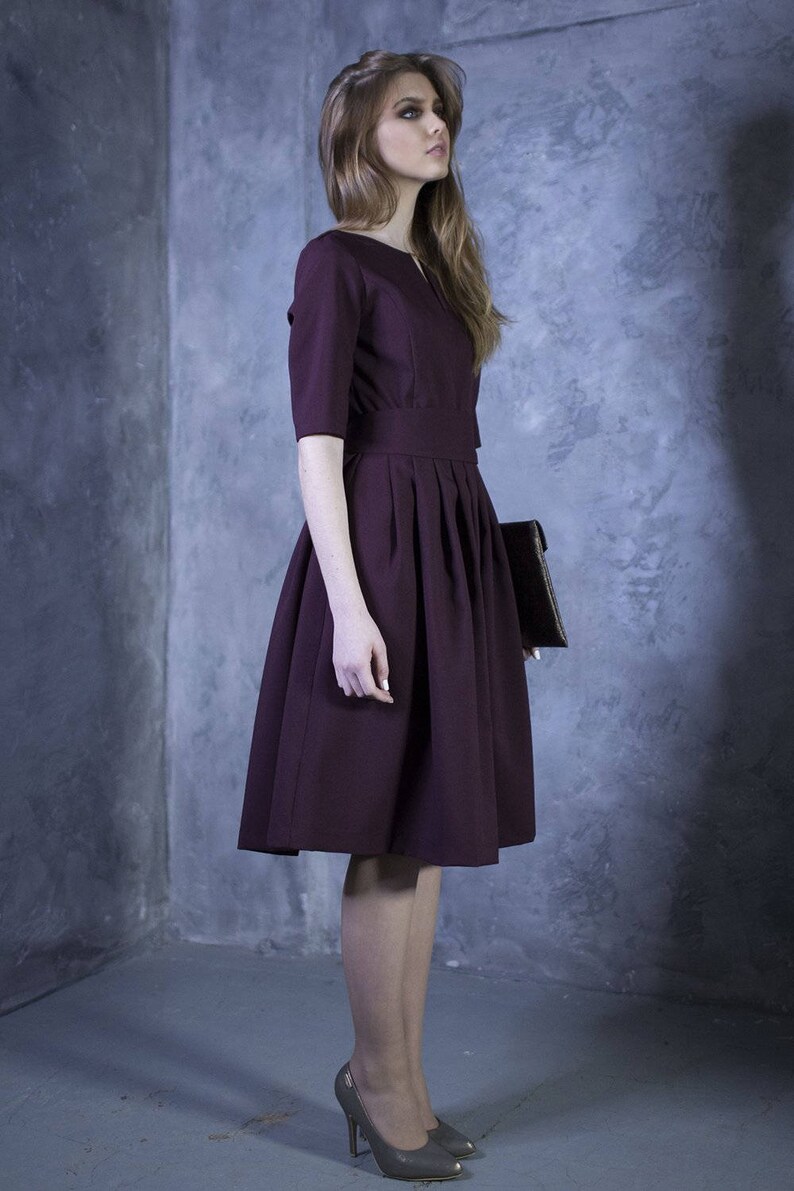 May include: A woman wearing a maroon dress with a pleated skirt and a brown clutch.