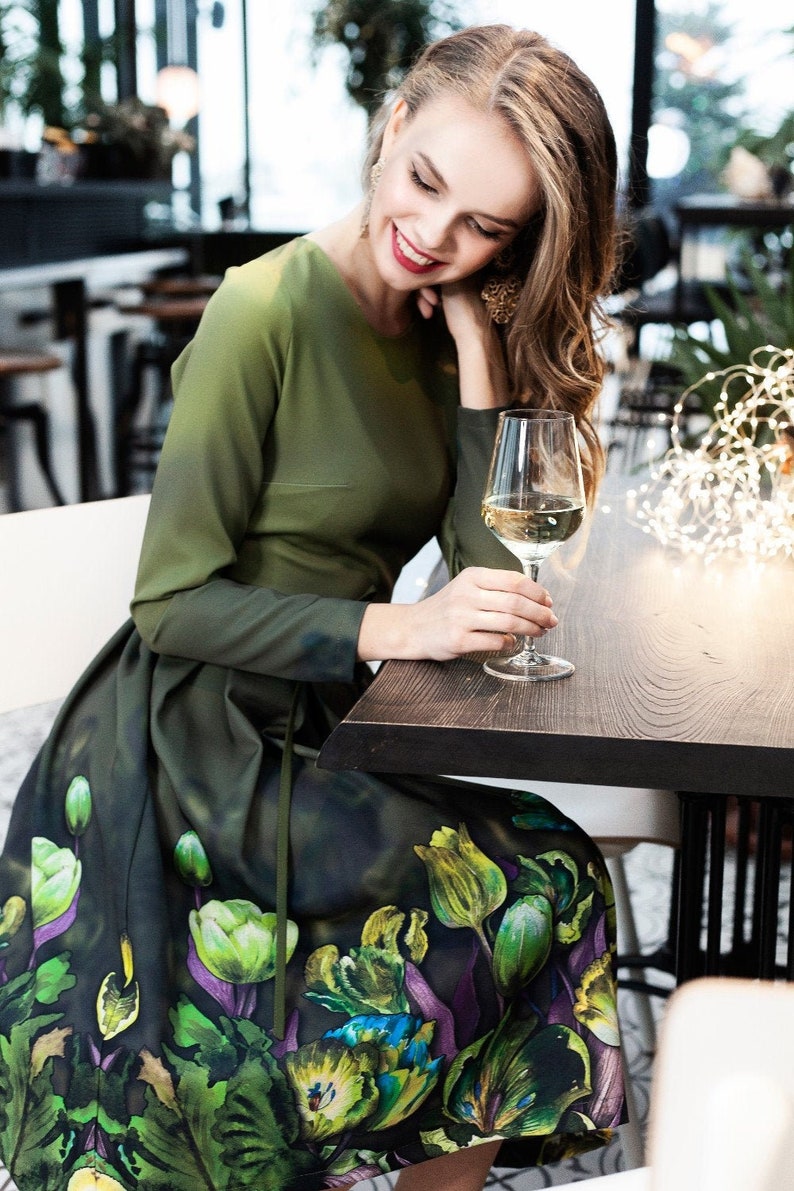 May include: A woman wearing a green dress with a floral print sits at a table with a glass of white wine. The dress has a green bodice and a skirt with a floral pattern in shades of green, purple, and yellow.