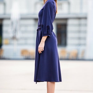 May include A woman wearing a dark blue midi dress with a belt and long sleeves. She is wearing burgundy ankle boots with a pointed toe.