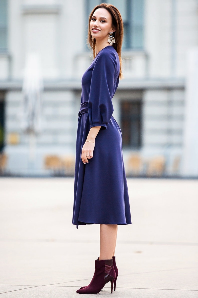 May include A woman wearing a dark blue midi dress with a belt and long sleeves. She is wearing burgundy ankle boots with a pointed toe.