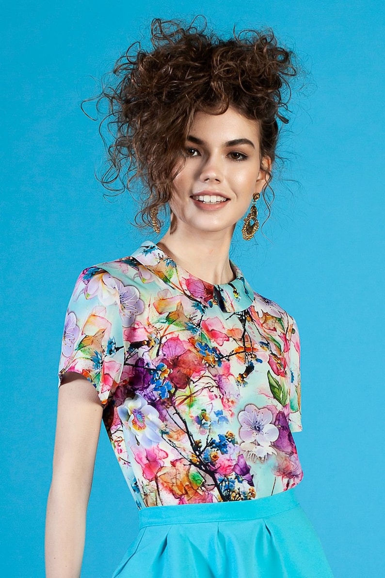 May include: A woman with curly brown hair is wearing a white blouse with a floral print and a turquoise skirt. The blouse has a Peter Pan collar and short sleeves.