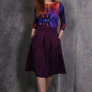 Pu&ograve; includere: Una donna indossa una gonna viola con una blusa con stampa astratta colorata e stivaletti marroni.