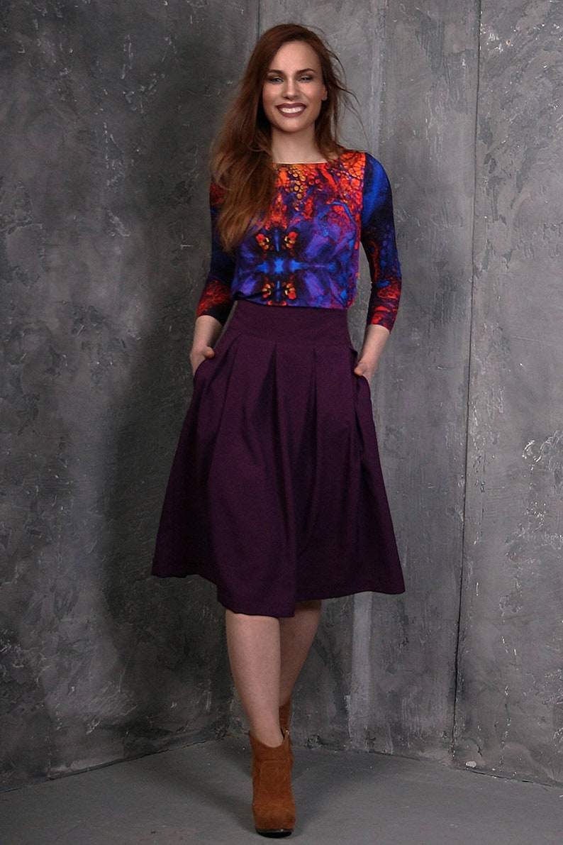 Pu&ograve; includere: Una donna indossa una gonna viola con una blusa con stampa astratta colorata e stivaletti marroni.