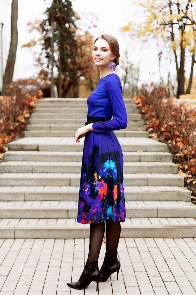 K&ouml;nnte beinhalten: Eine Frau in einem blauen Kleid mit einem Rock mit Blumenmuster und schwarzen Stiefeletten mit Absatz. Sie steht auf einer Treppe mit Herbstlaub im Hintergrund.