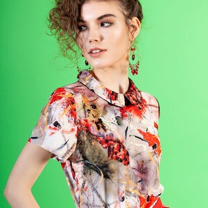 May include: A woman with curly brown hair is wearing a white blouse with a floral print and a red skirt. She is wearing red earrings.