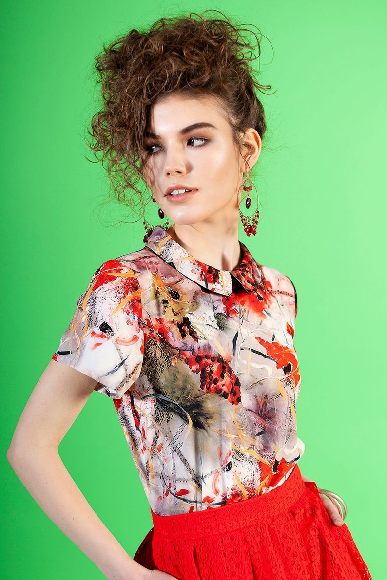 May include: A woman with curly brown hair is wearing a white blouse with a floral print and a red skirt. She is wearing red earrings.