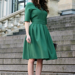 May include: A woman wearing a green knee-length dress with a fitted bodice and a full skirt. She is standing on a set of stairs.