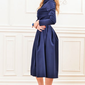Pu&ograve; includere: Un abito midi blu navy con maniche lunghe e chiusura con bottoni sul davanti. L'abito ha un corpetto aderente e una gonna ampia con pieghe. La modella indossa tacchi alti neri.