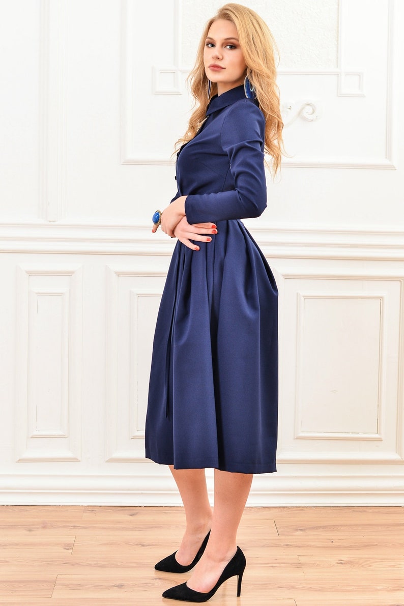 Pu&ograve; includere: Un abito midi blu navy con maniche lunghe e chiusura con bottoni sul davanti. L'abito ha un corpetto aderente e una gonna ampia con pieghe. La modella indossa tacchi alti neri.