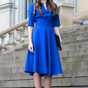 May include: A royal blue dress with a v-neckline and a fitted bodice. The skirt is full and falls to the knee. The dress is worn by a model standing on a set of stairs.