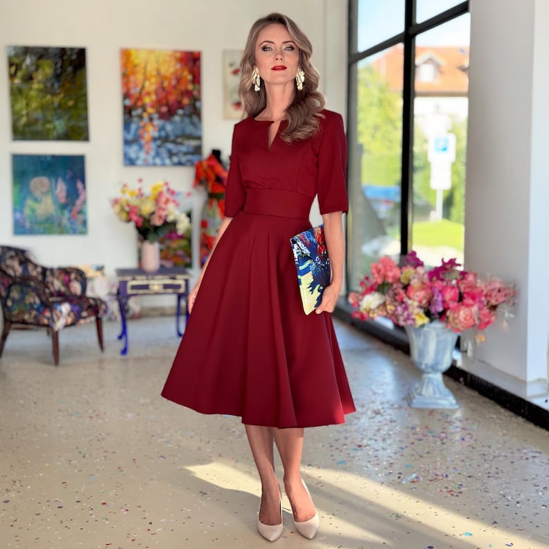 Op de afbeelding: Een vrouw in een bordeauxrode midi-jurk met een v-hals en een getailleerde taille. Ze houdt een kleurrijke clutch vast en staat in een kamer met kunstwerken aan de muren.