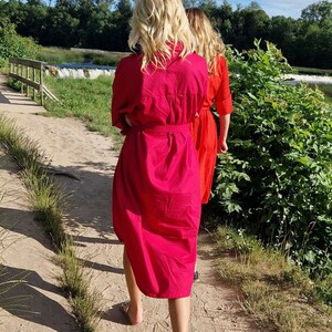 May include: Two women walking on a sandy path, wearing bright, solid-colored dresses. The woman in front is wearing a fuchsia dress with a matching belt, while the woman behind her is wearing a red dress. The background includes a waterfall and lush greenery.