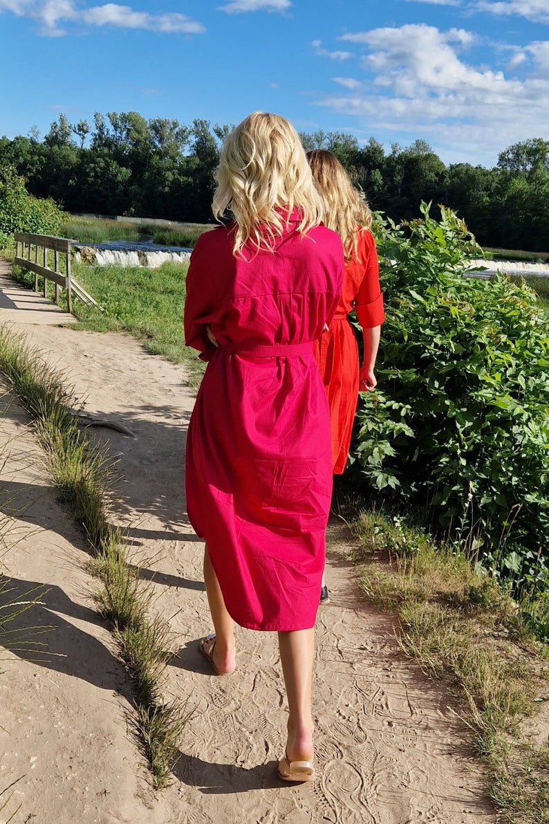 May include: Two women walking on a sandy path, wearing bright, solid-colored dresses. The woman in front is wearing a fuchsia dress with a matching belt, while the woman behind her is wearing a red dress. The background includes a waterfall and lush greenery.