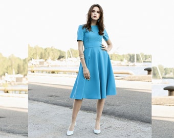 Pode incluir: Uma mulher vestindo um vestido azul claro até o joelho com um corpete justo e uma saia cheia. Ela está em pé em uma superfície pavimentada com um fundo desfocado.