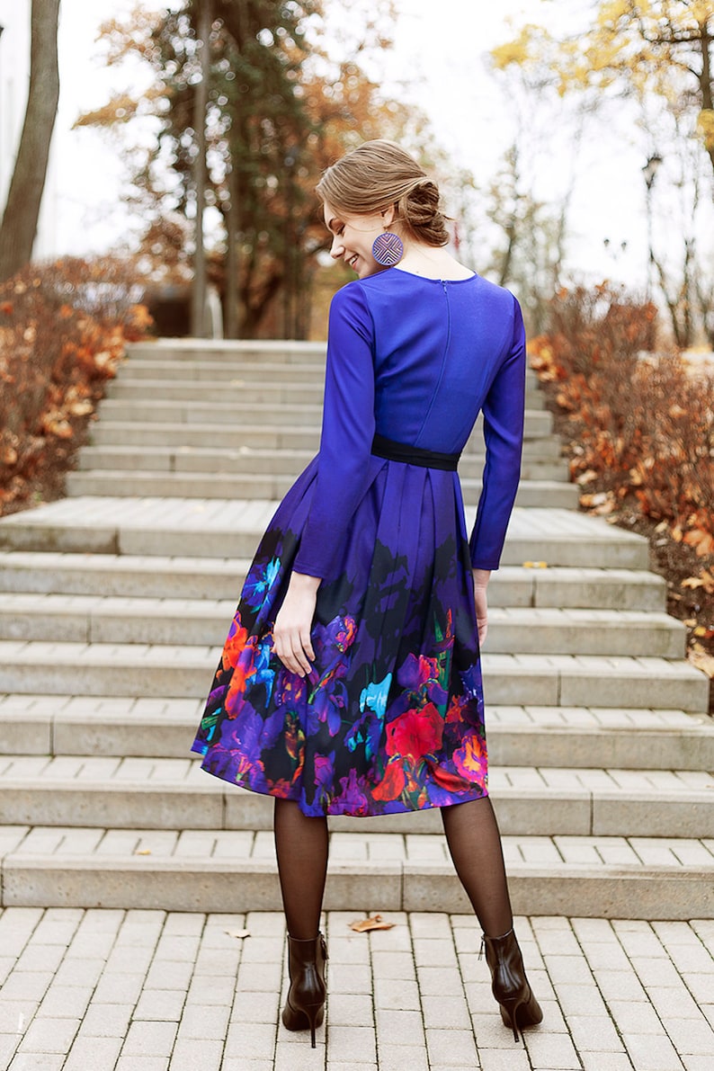 K&ouml;nnte beinhalten: Eine Frau in einem lila Kleid mit Blumenmuster und schwarzen Strumpfhosen. Sie steht auf einer Treppe mit Herbstlaub im Hintergrund.