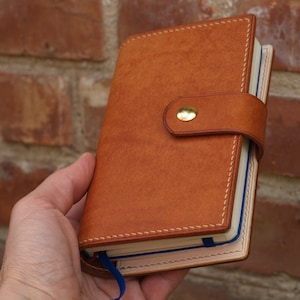 Peut inclure: Une couverture de carnet en cuir marron avec une fermeture à bouton doré et un élastique bleu. La couverture a un bord cousu et un intérieur crème.