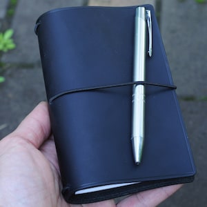 May include: A black leather notebook with a silver pen secured with an elastic band. The notebook is closed and has a plain cover.