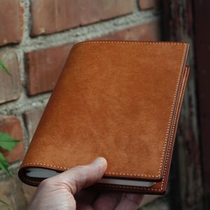 May include: A tan leather journal with a stitched border. The journal is held open, showing the white pages within. The leather has a natural grain and a warm colour. The stitching is a lighter shade, providing contrast.