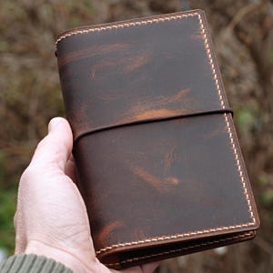 May include: A brown leather journal with a distressed finish, held in a hand. The journal is rectangular, with a stitched border and a dark elastic band closure. The leather has a mottled appearance with lighter brown highlights.