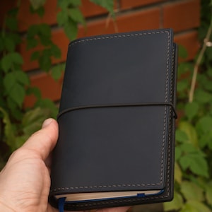 Può includere: Una copertina per quaderno in pelle blu scuro con dettagli di cucitura grigi e una chiusura a fascia elastica.